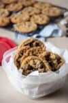 A plastic container with chocolate chip cookies in it with a rack of cooling cookies in the back.