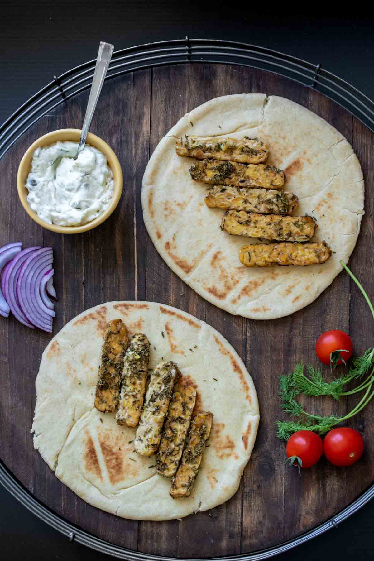 Slices of tempeh on two pita bread on a wooden surface and other ingredients for gyros around them.