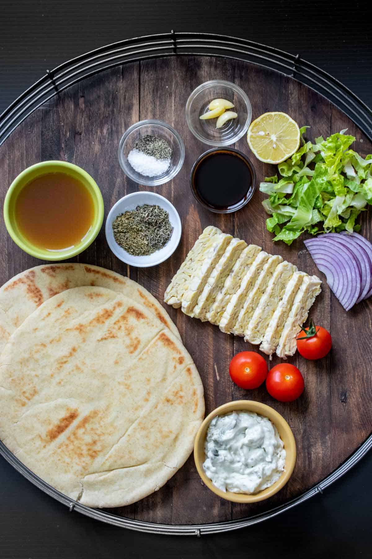 A wooden tray with metal handles and all the ingredients needed to marinate and build tempeh gyros.