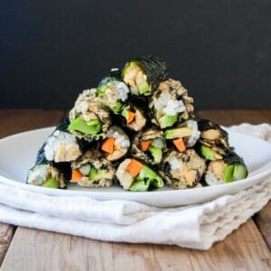 A white plate of a pile of veggie and rice filled seaweed snacks on a white towel sitting on a wooden surface.