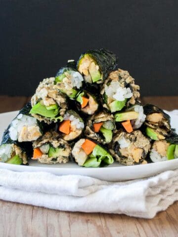 A white plate of a pile of veggie and rice filled seaweed snacks on a white towel sitting on a wooden surface.