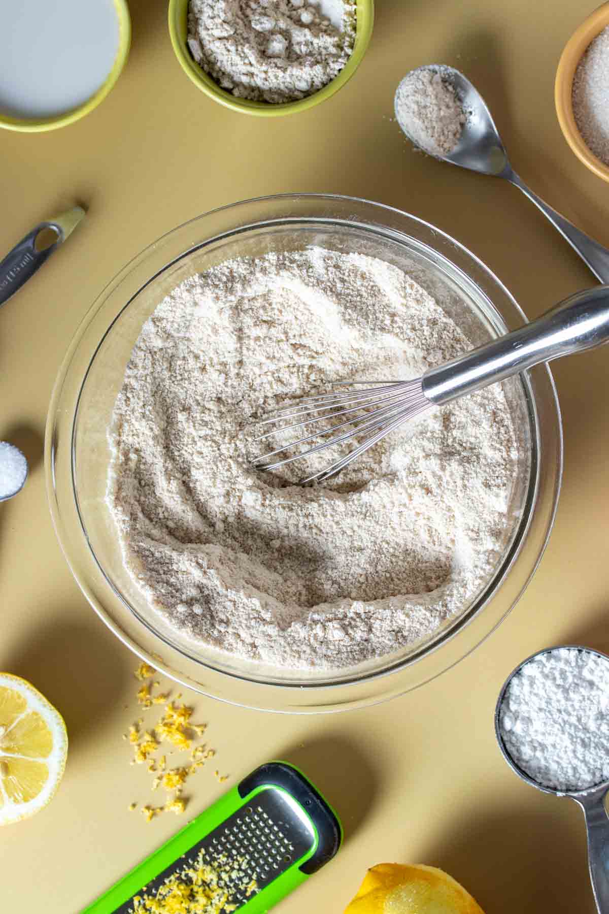 A whisk mixing flours in a glass bowl sitting on a yellow surface.