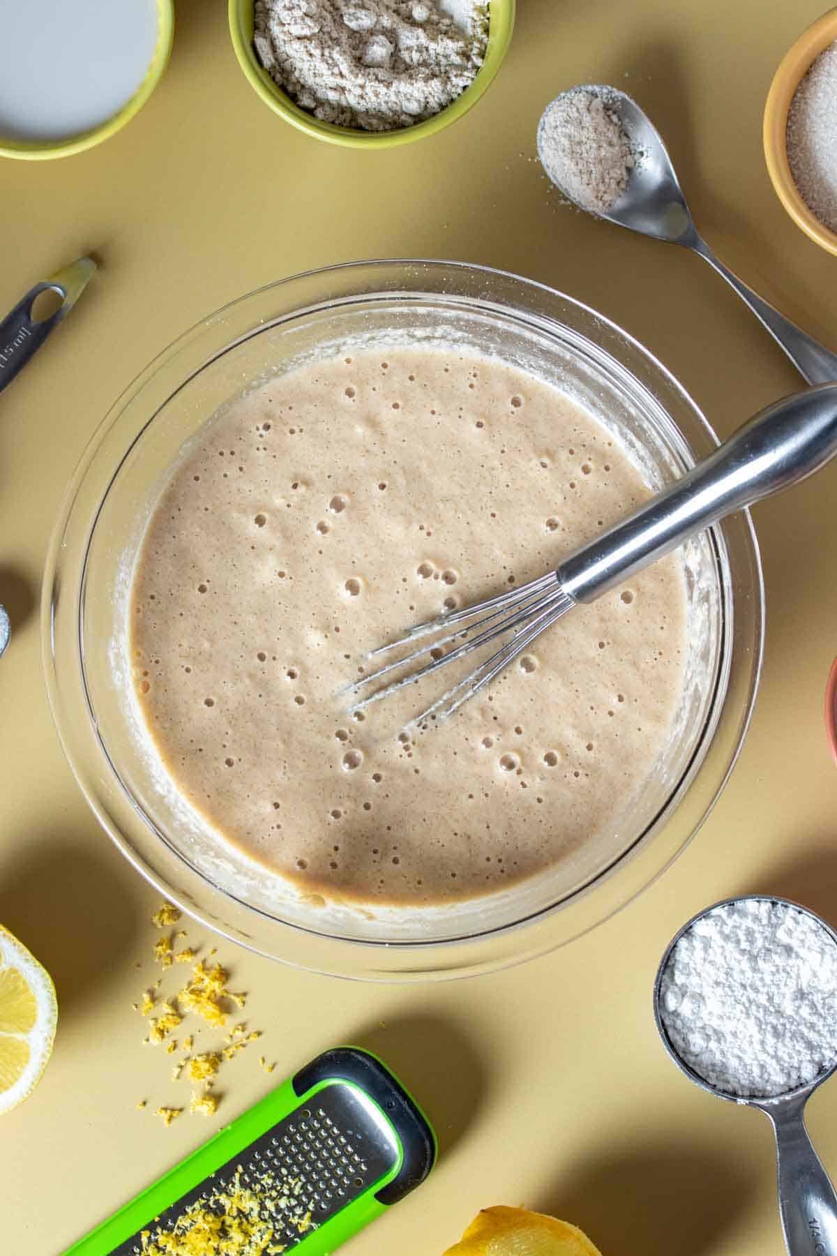 A whisk in a glass bowl filled with tan colored batter on a yellow surface.