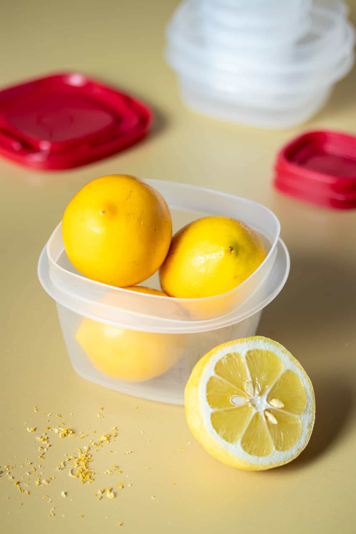 A plastic container filled with lemons and a half lemon in front of it with more containers in the background.