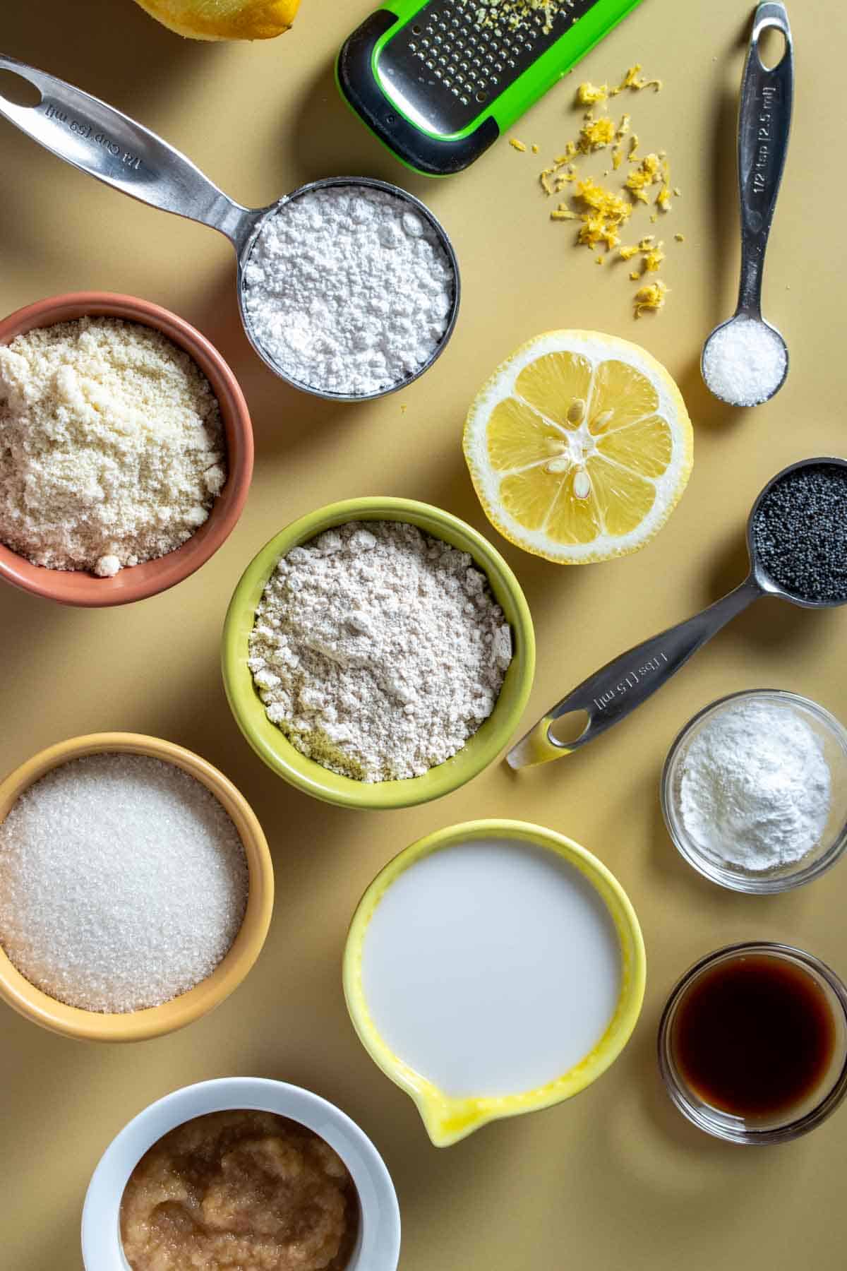 Top view of ingredients needed to make lemon poppy seed muffins, some in colored bowls and some right on the yellow surface.