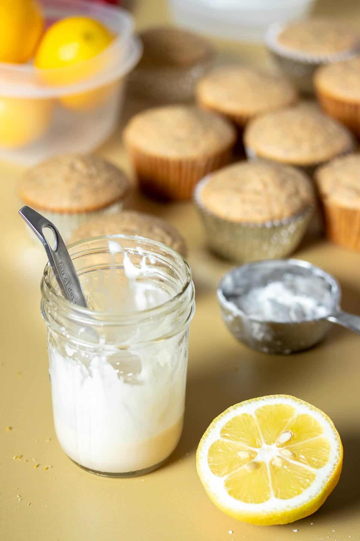 A glass jar with some white glaze and a half lemon next to it in front of a group of muffins.