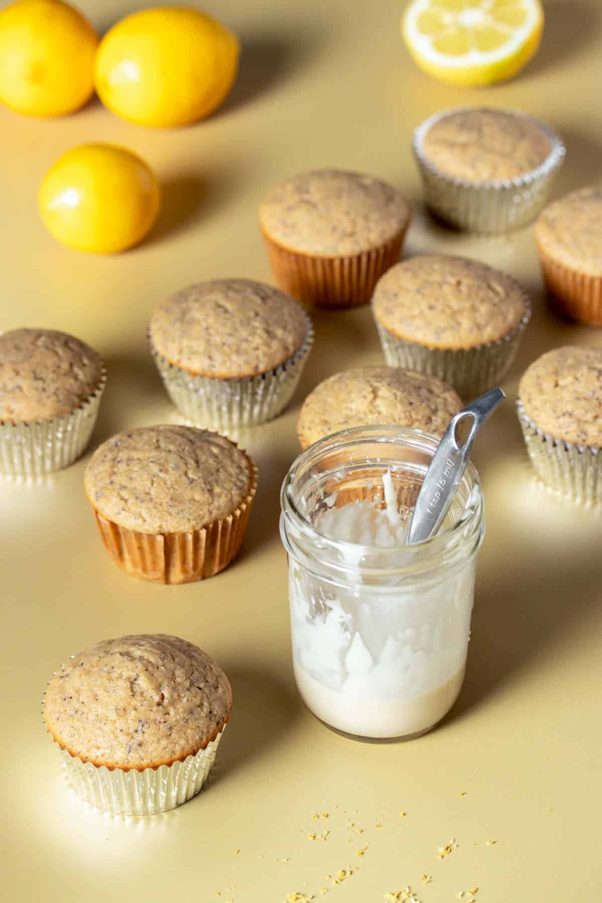 A group of lemon poppy seed muffins without glaze surrounding a glass jar with white glaze in it.