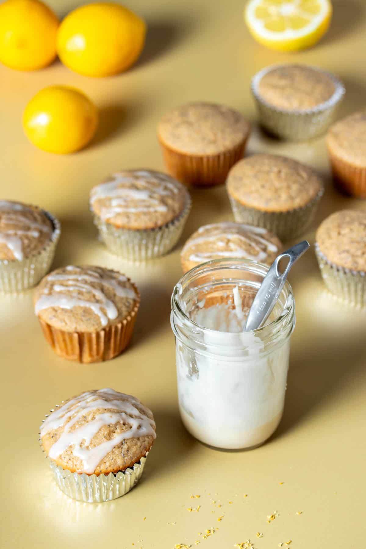 A glass jar of white glaze next to lemon poppy seed muffins that have just been glazed.