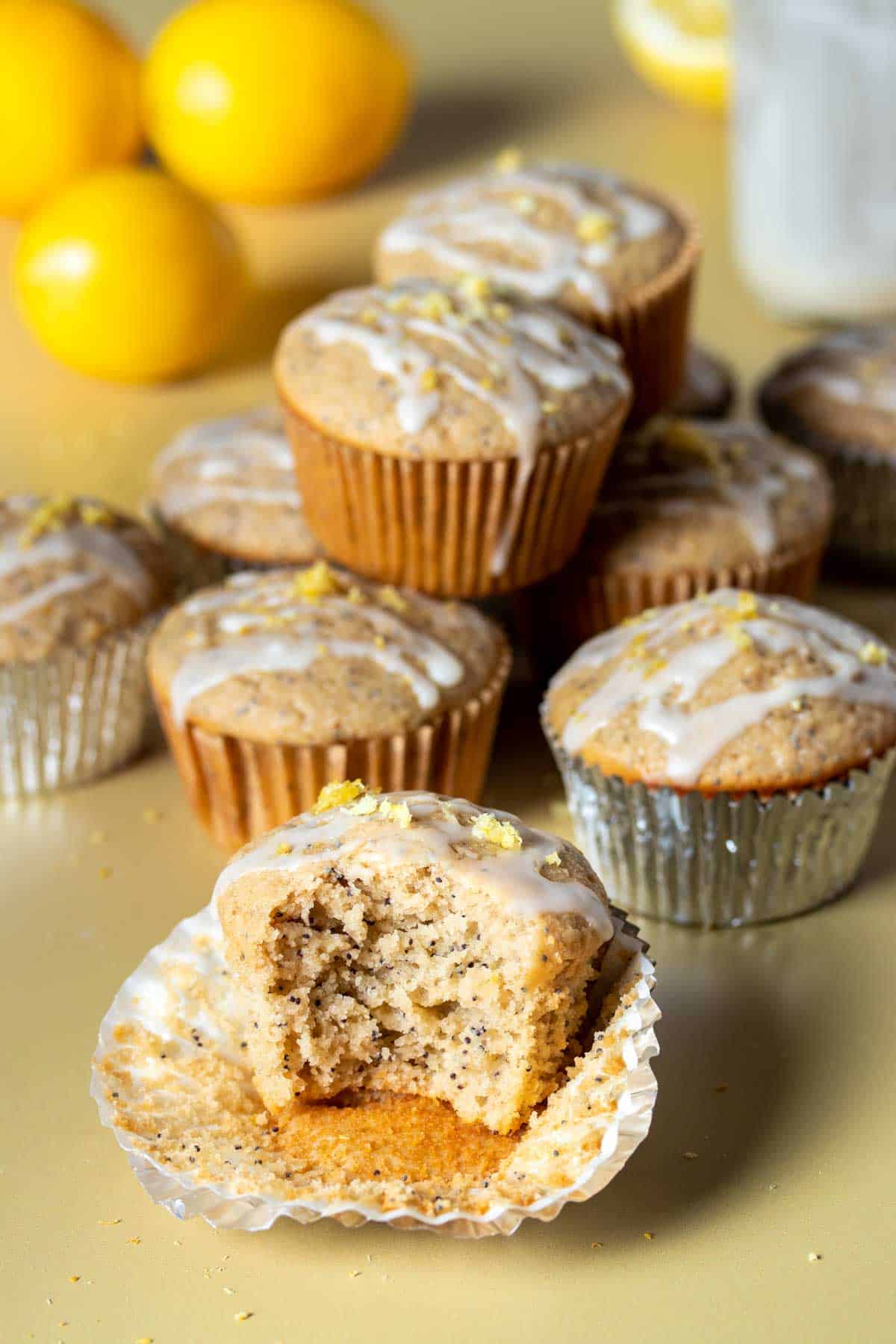 A group of lemon poppy seed muffins in front of lemons and one with a bite out of it in the front.