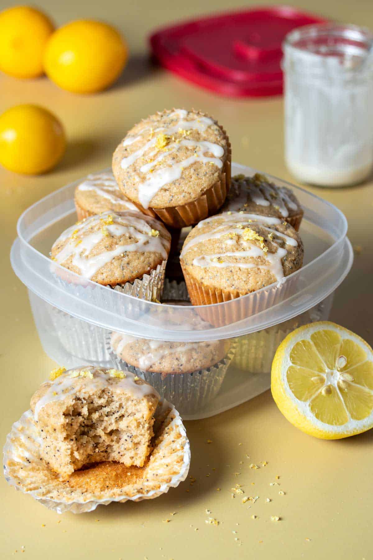 A plastic container filled with lemon poppy seed muffins and one with a bite out of it sitting in front.