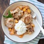 A white bowl with brown rim filled with peach cobbler topped with whipped cream and a fork in it.