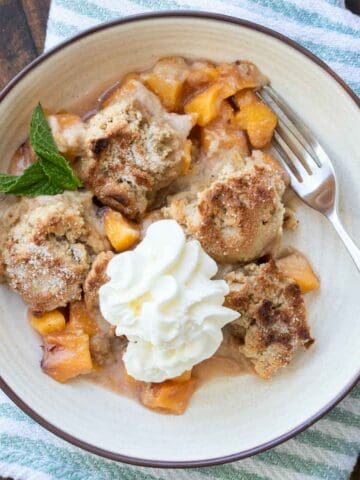 A white bowl with brown rim filled with peach cobbler topped with whipped cream and a fork in it.