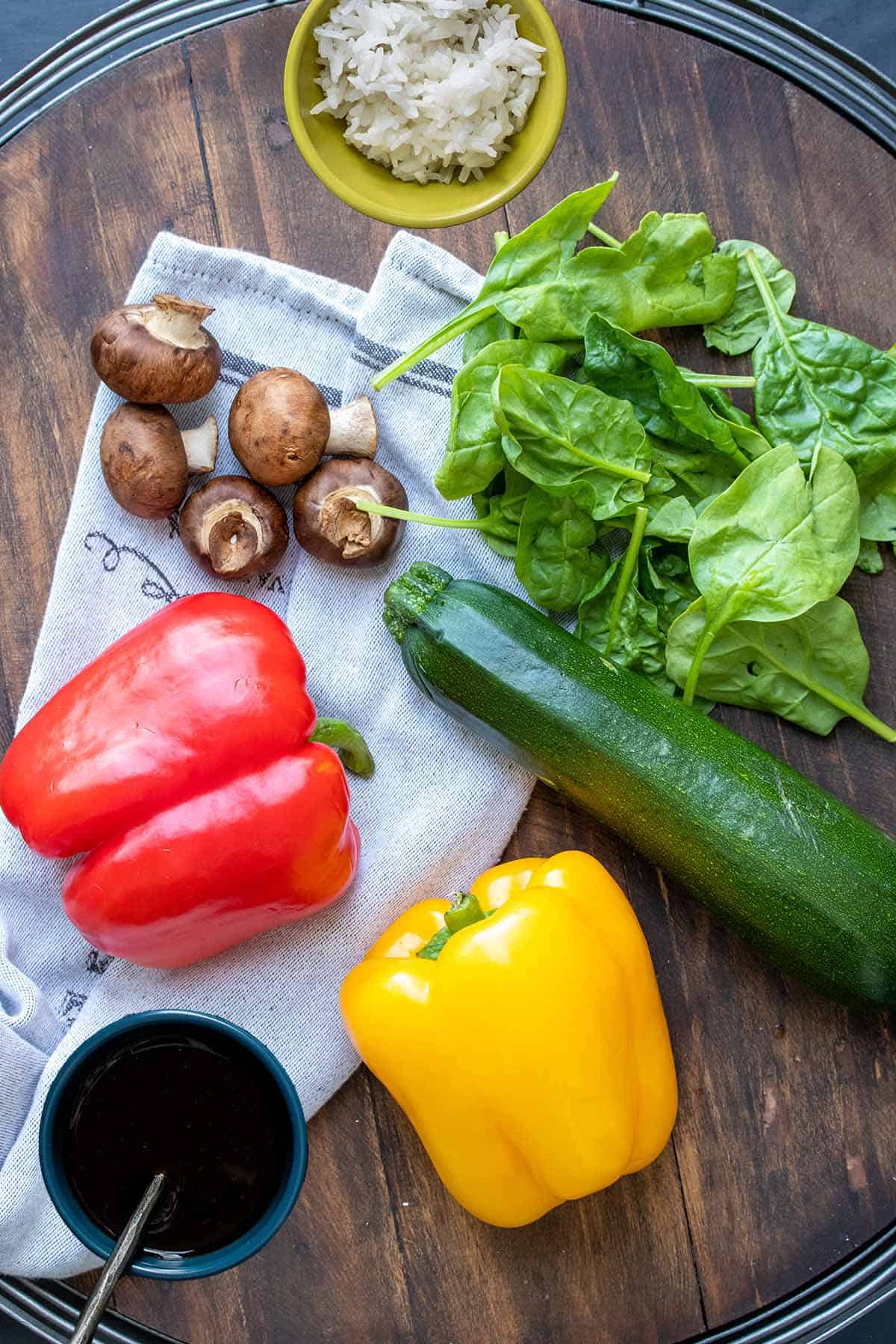 A wooden round tray with peppers, spinach, mushrooms and zucchini on it next to a bowl of white rice and teriyaki sauce.