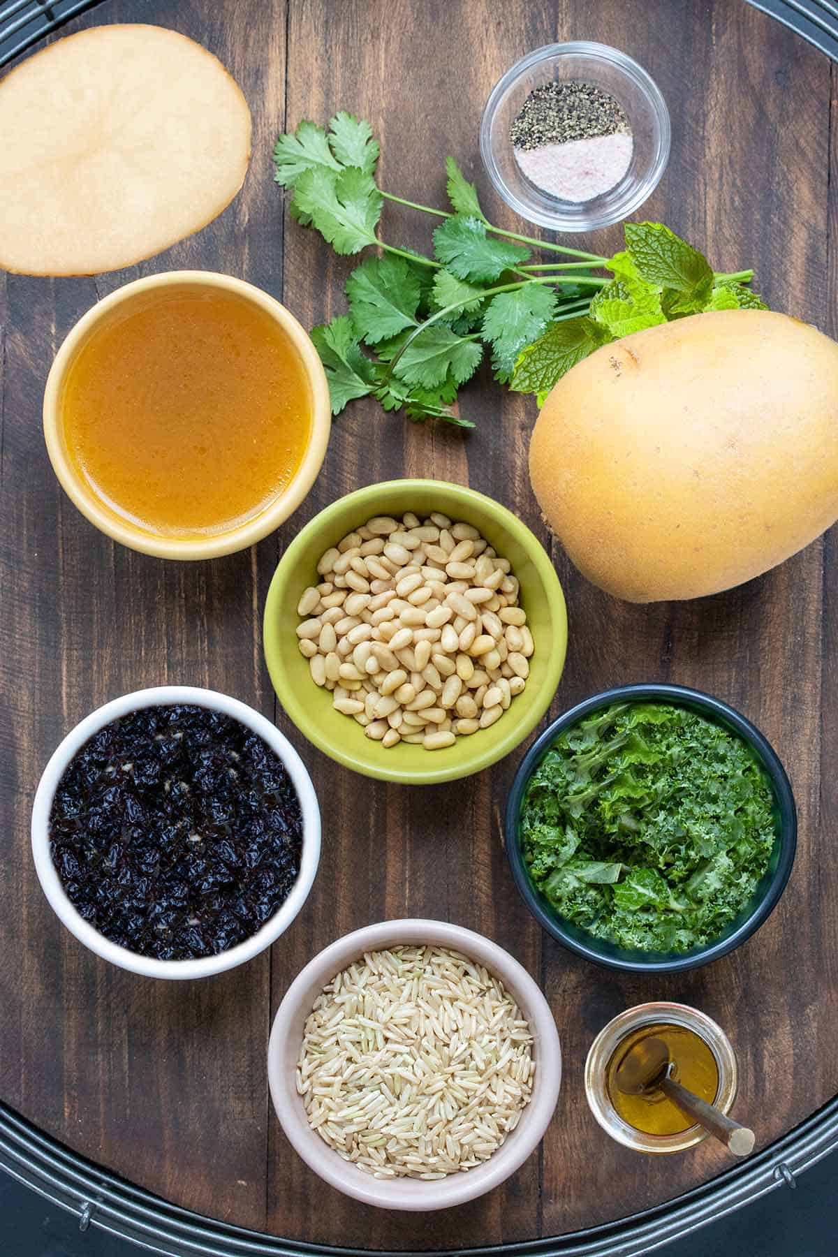 Top view of bowls with broth, nuts, kale, rice, currants, salt and pepper and oil next to parsley and potatoes.