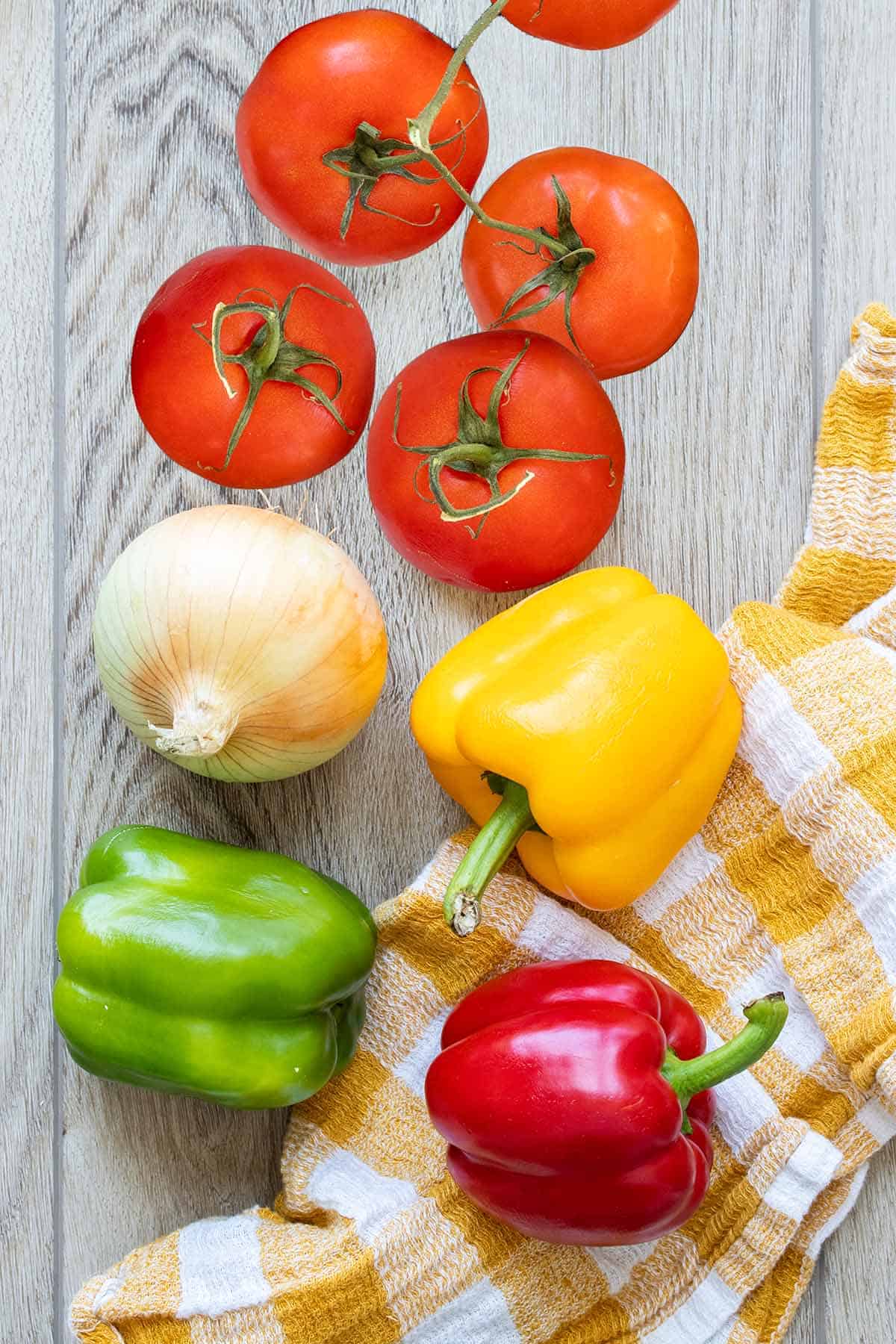 Top view of fresh tomatoes, peppers and an onion on a tan surface with a yellow checked towel.