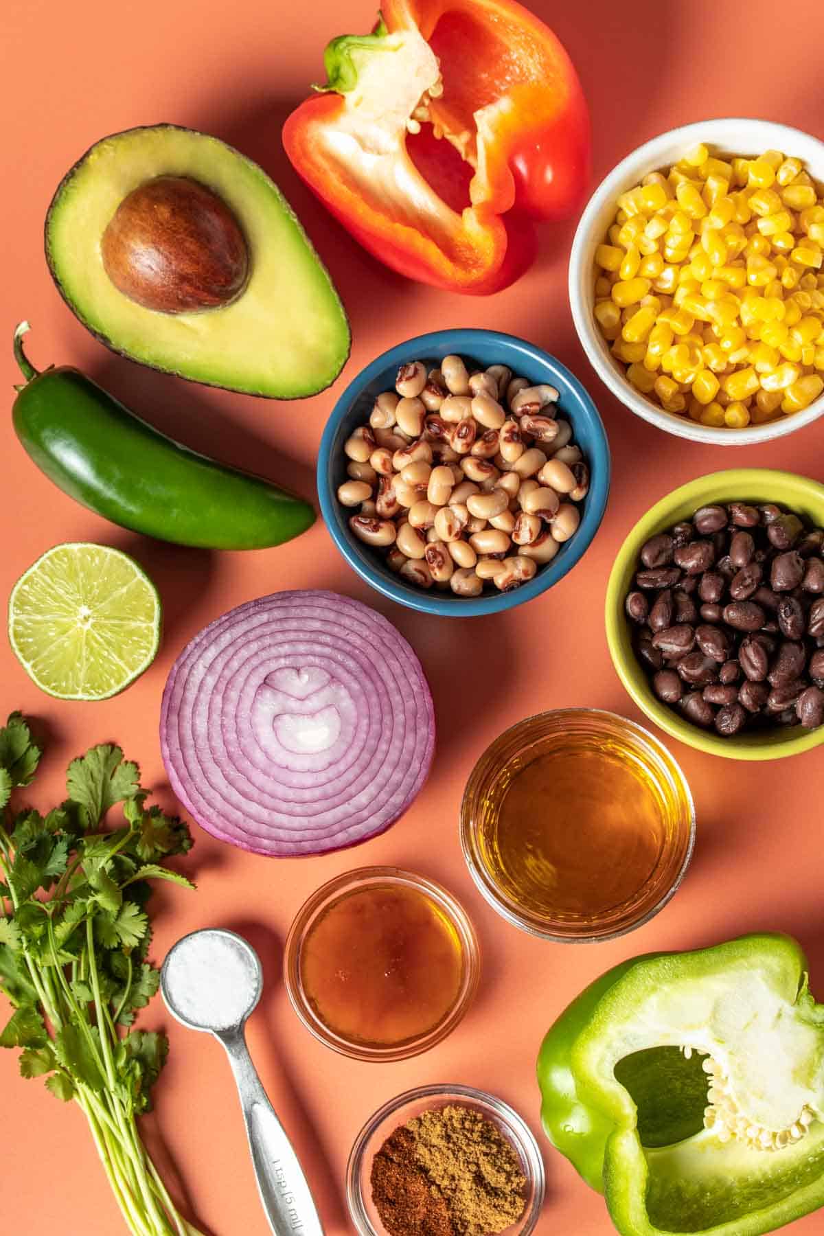 Bowls of beans, corn and dressing ingredients with half cut lime, avocado, onion and peppers and sprigs of cilantro on an orange background.