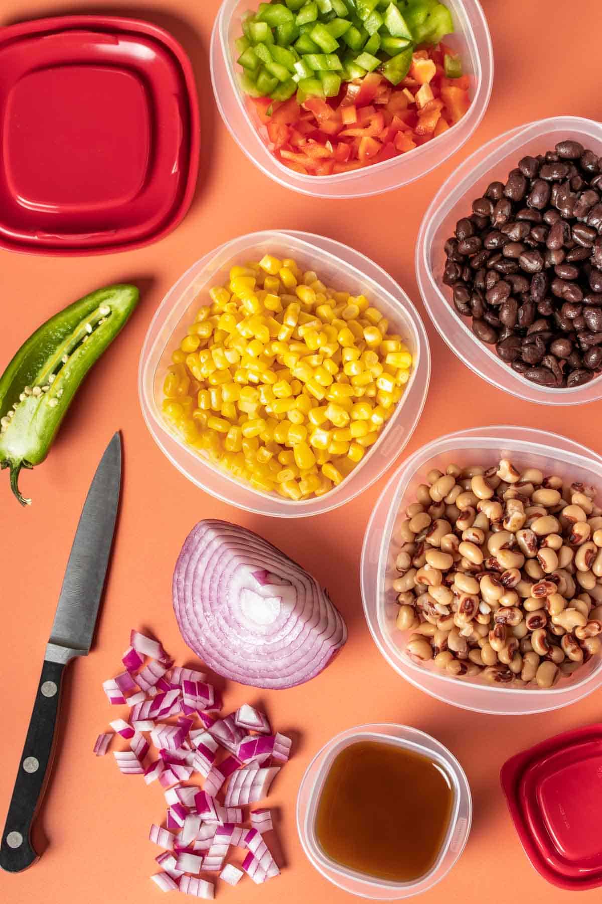 Prepped ingredients for a cowboy caviar salad in plastic containers sitting on an orange background.