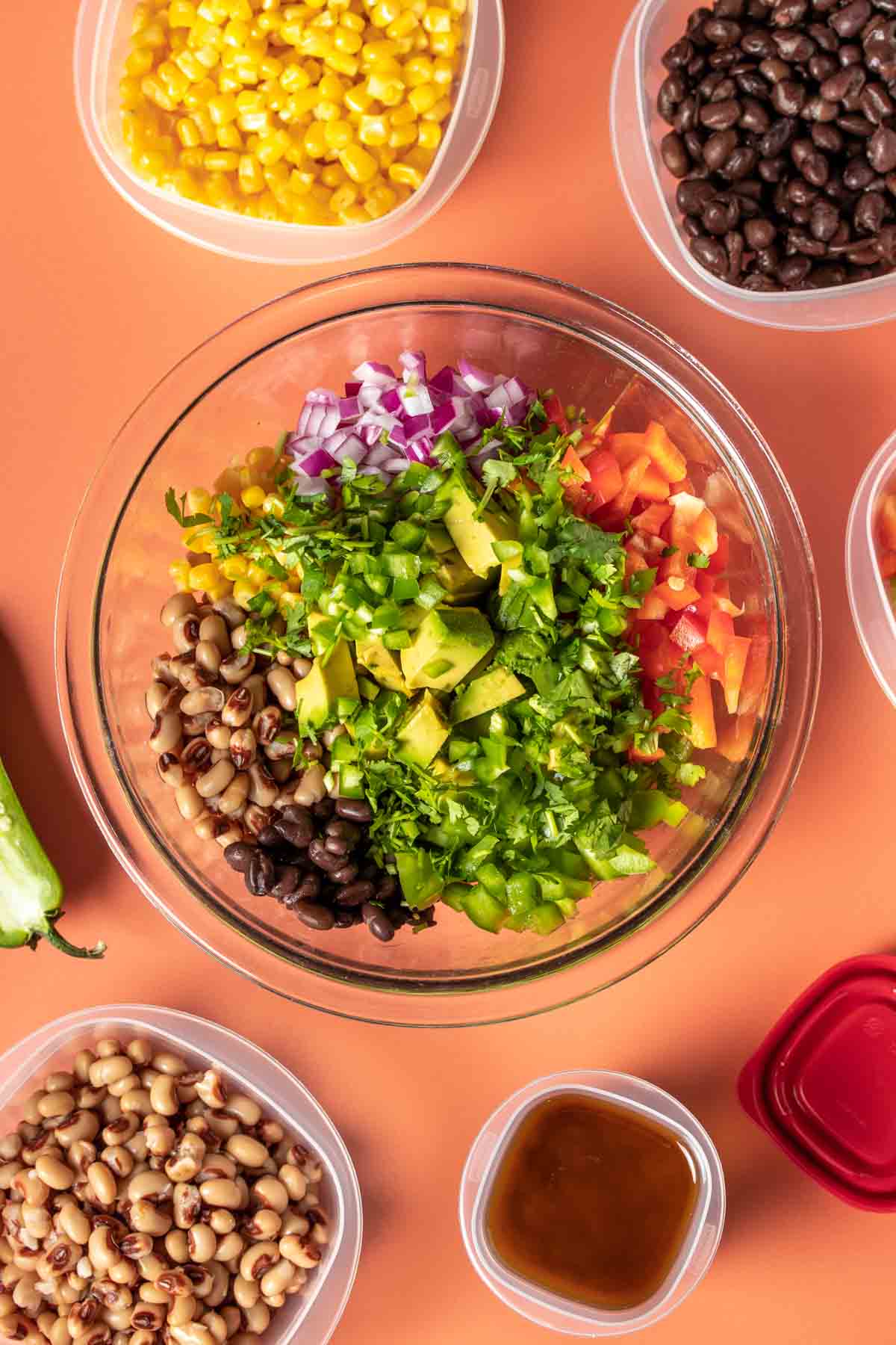A glass bowl with ingredients to make cowboy caviar lined up surrounded by more ingredients in bowls on an orange background.