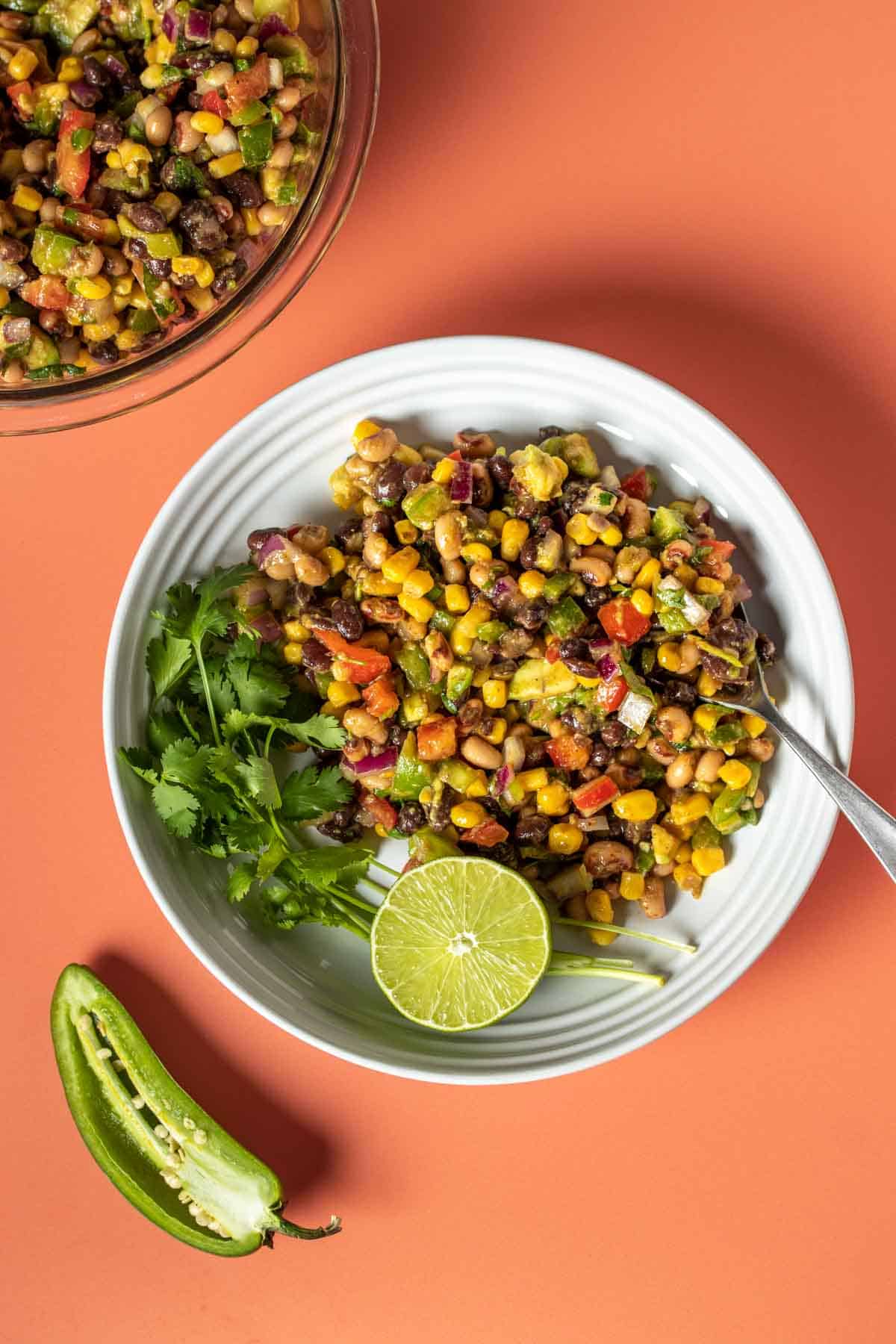 An orange background with a white bowl filled with a cowboy caviar salad next to sprigs of cilantro and half a lime sitting next to half a jalapeno.