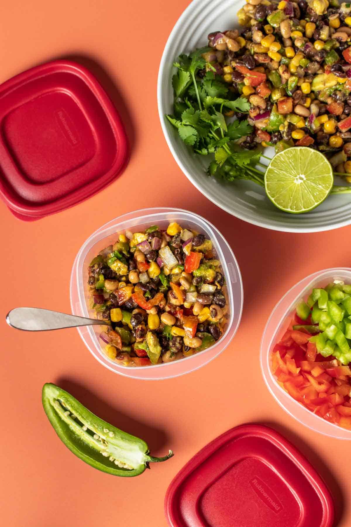 A plastic container with cowboy caviar in it next to a bowl of more salad and a plastic container with chopped peppers.