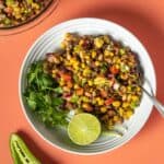 A cowboy caviar salad in a white bowl with springs of cilantro and half a lime sitting on an orange background.