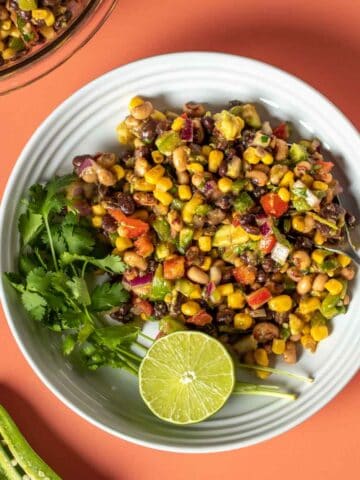 A cowboy caviar salad in a white bowl with springs of cilantro and half a lime sitting on an orange background.