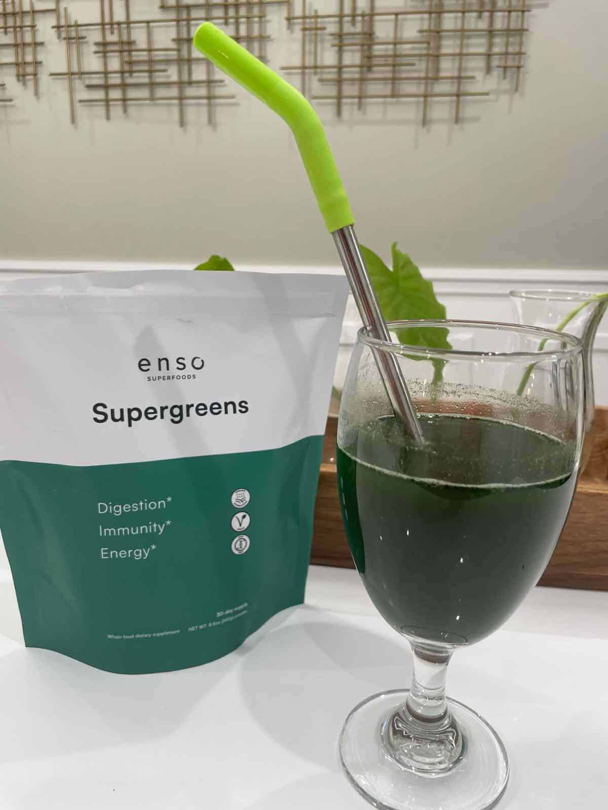 A glass of green juice on a counter next to a green and white bag of greens powder.