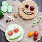 Rice cakes with foods used to make faces on them on a piece of parchment next to cucumber, tomatoes and nuts.