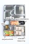 A white background with a metal lunch box with sections that have veggies, fruit, snacks and tortilla roll ups.