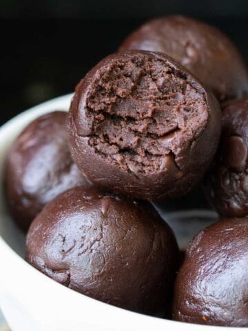 A white bowl with a pile of round brownie bites in it and one on the top has a bite out of it.