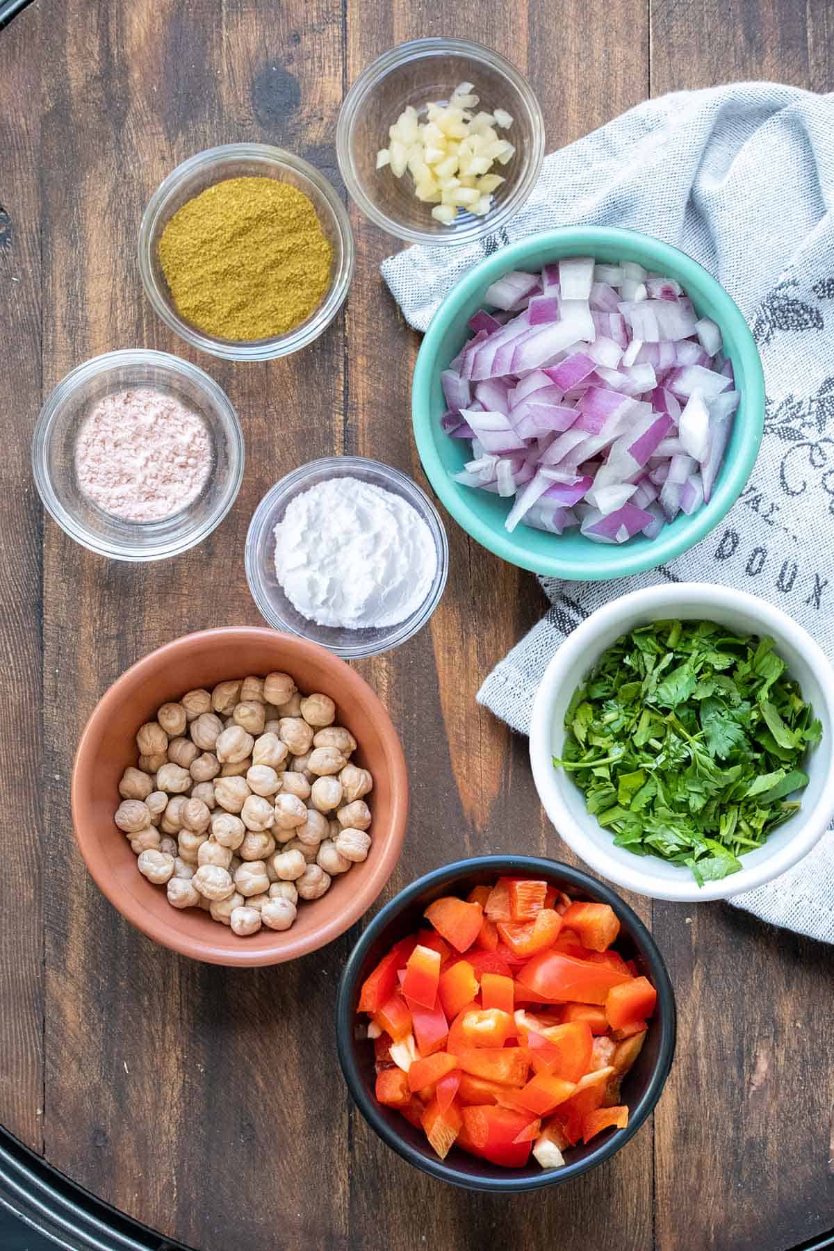 Top view of colorful and glass bowls with ingredients needed to make a falafel burger on a wooden surface.