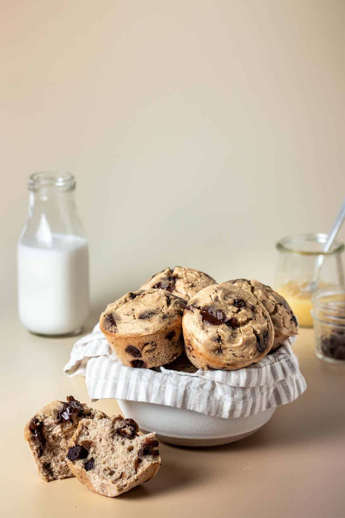 A muffin opened up in half in front of a white bowl with a tan striped towel filled with other muffins on a tan surface.