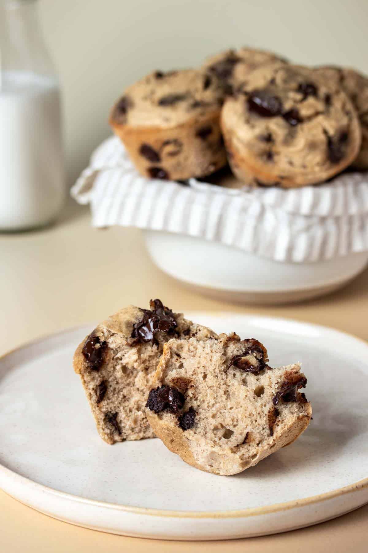 A muffin cut in half laying on a tan plate in front of a bowl of muffins and a jar of milk.