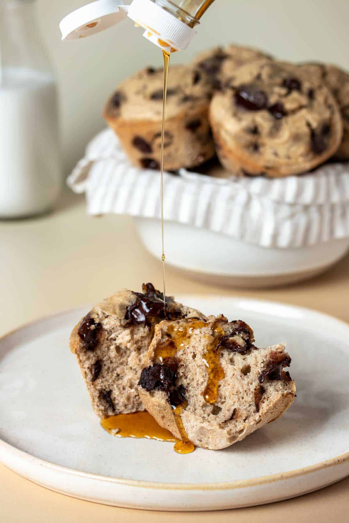 Maple syrup drizzling on a muffin cut in half laying on a tan plate in front of a white bowl with a towel filled with muffins.