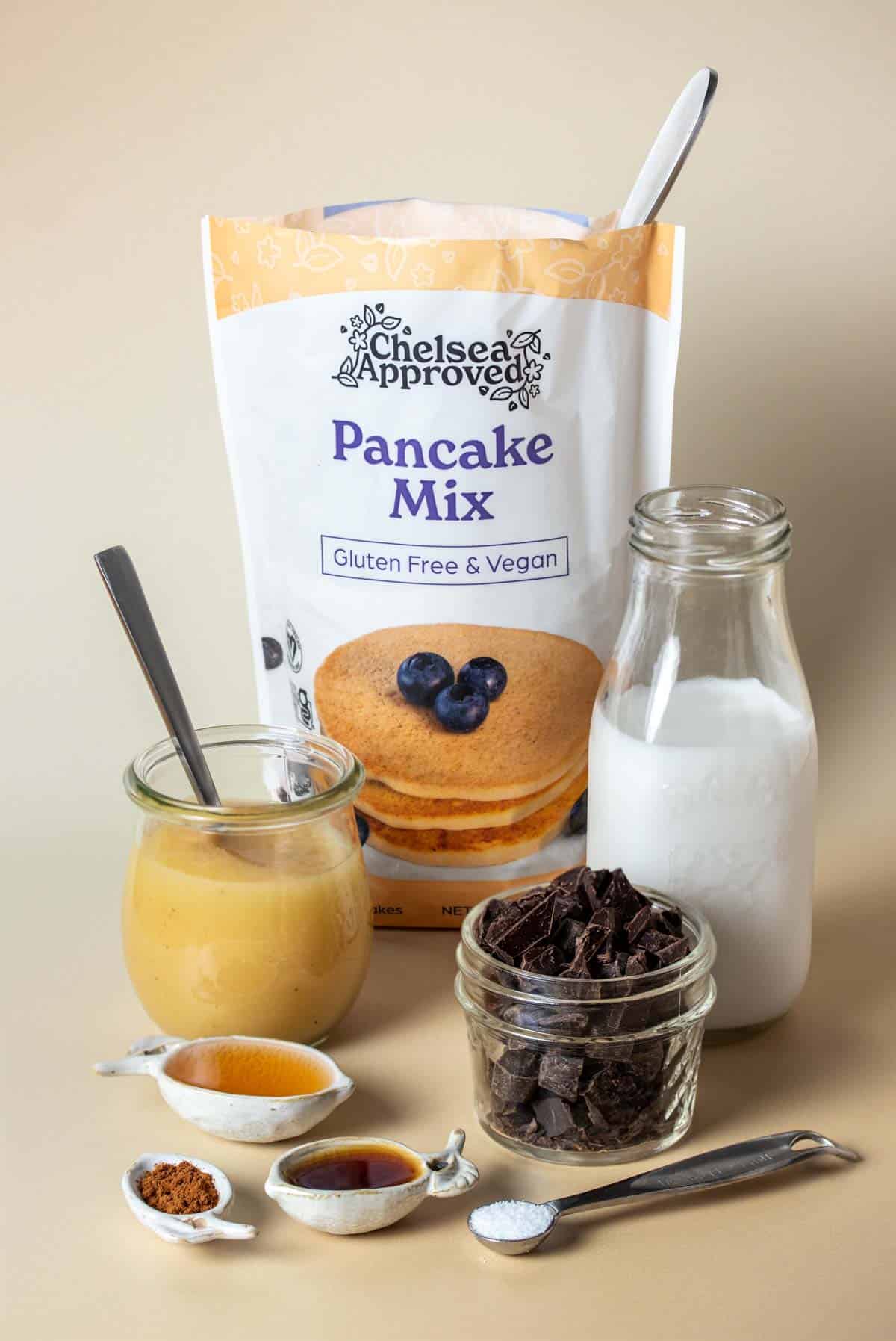 A bag of pancake mix on a tan surface with apple sauce, milk, chocolate chips, seasonings, and brown liquids in different bowls and spoons.