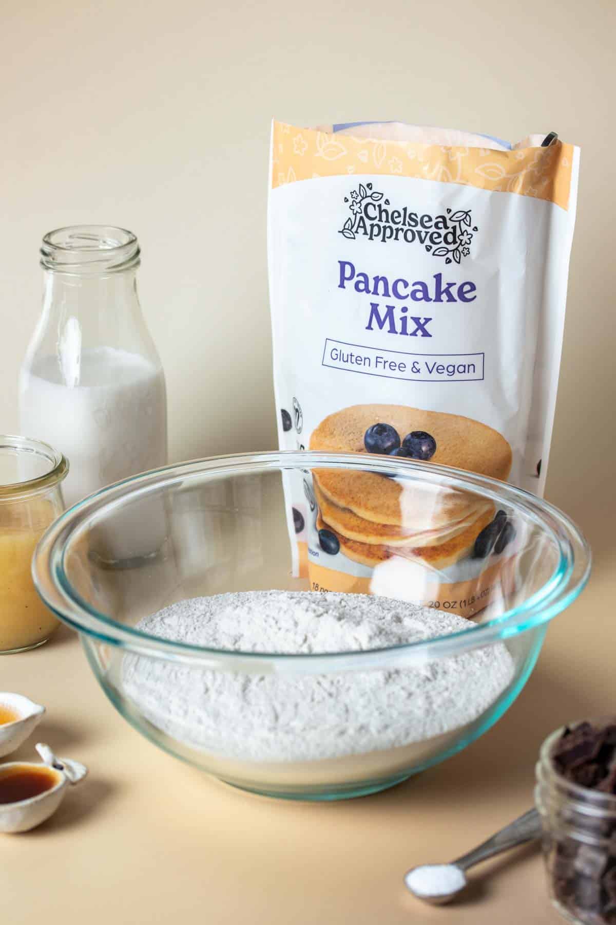 A glass bowl filled with flour in front of a bag of pancake mix surrounded by ingredients to make pancakes.