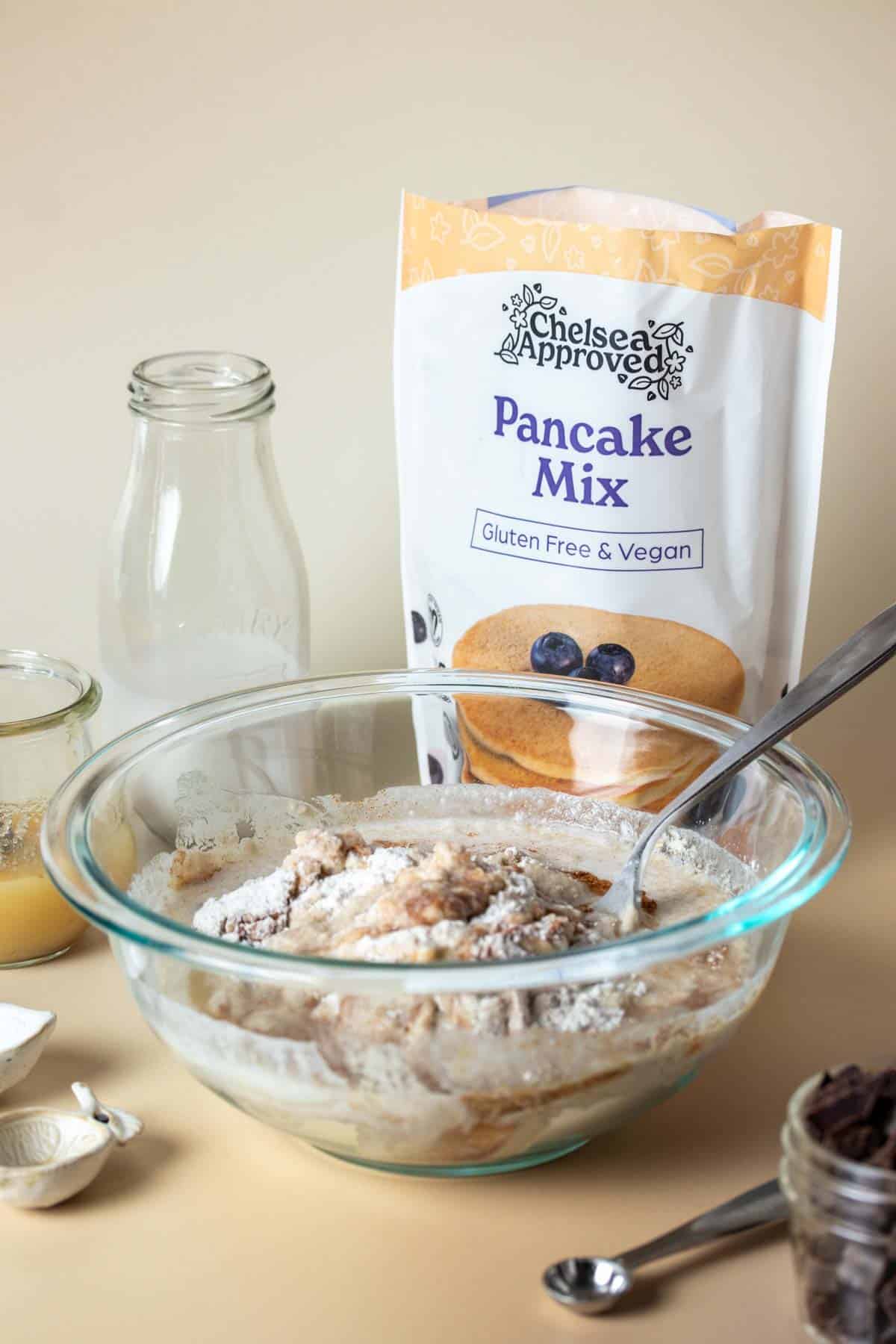 A glass bowl with ingredients to make pancake muffins about to be mixed together on a tan surface surrounded by more of those other ingredients.