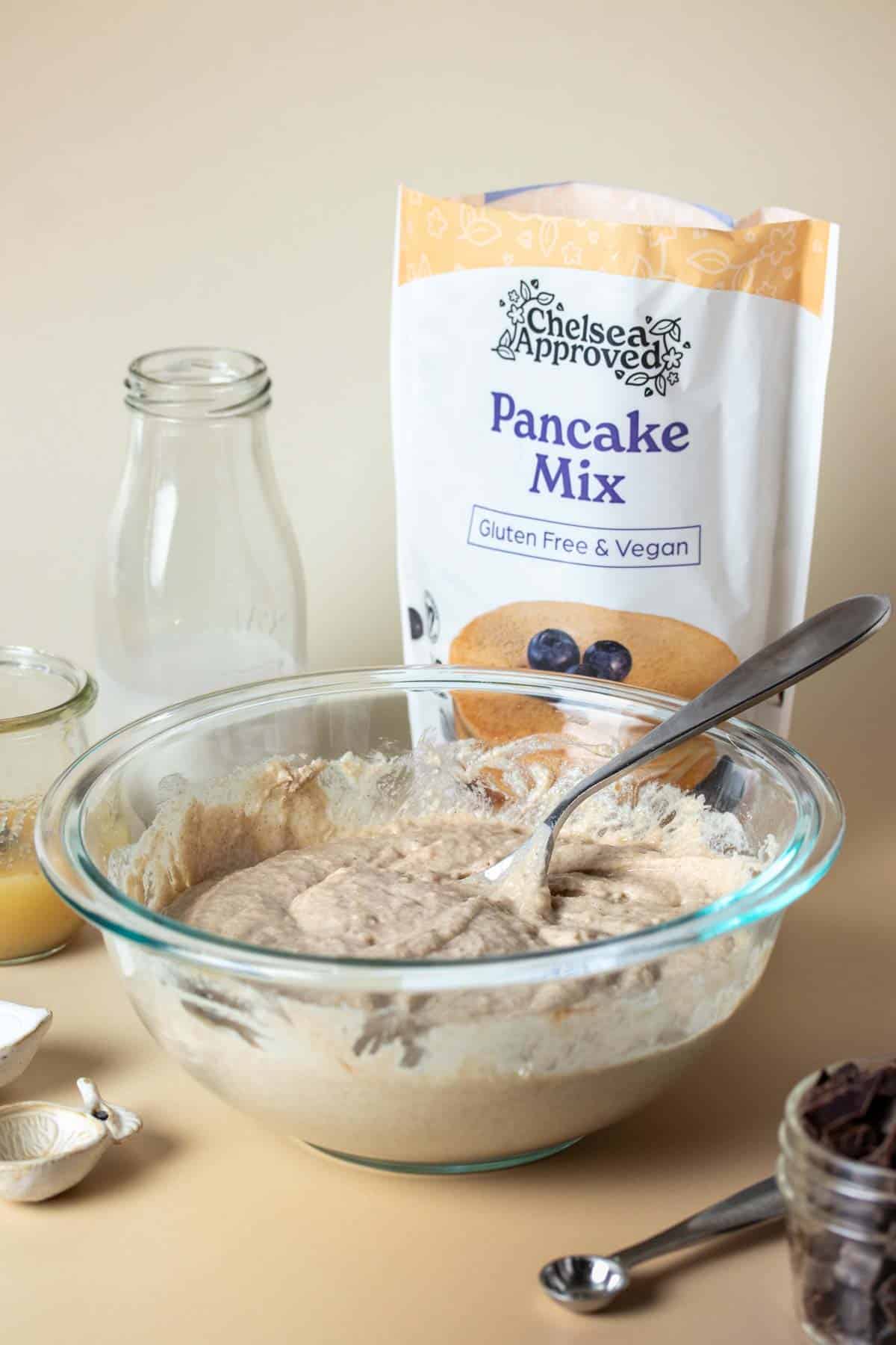 A pancake muffin batter in a glass bowl with a spoon in it in front of a bag of pancake mix and other ingredients used.