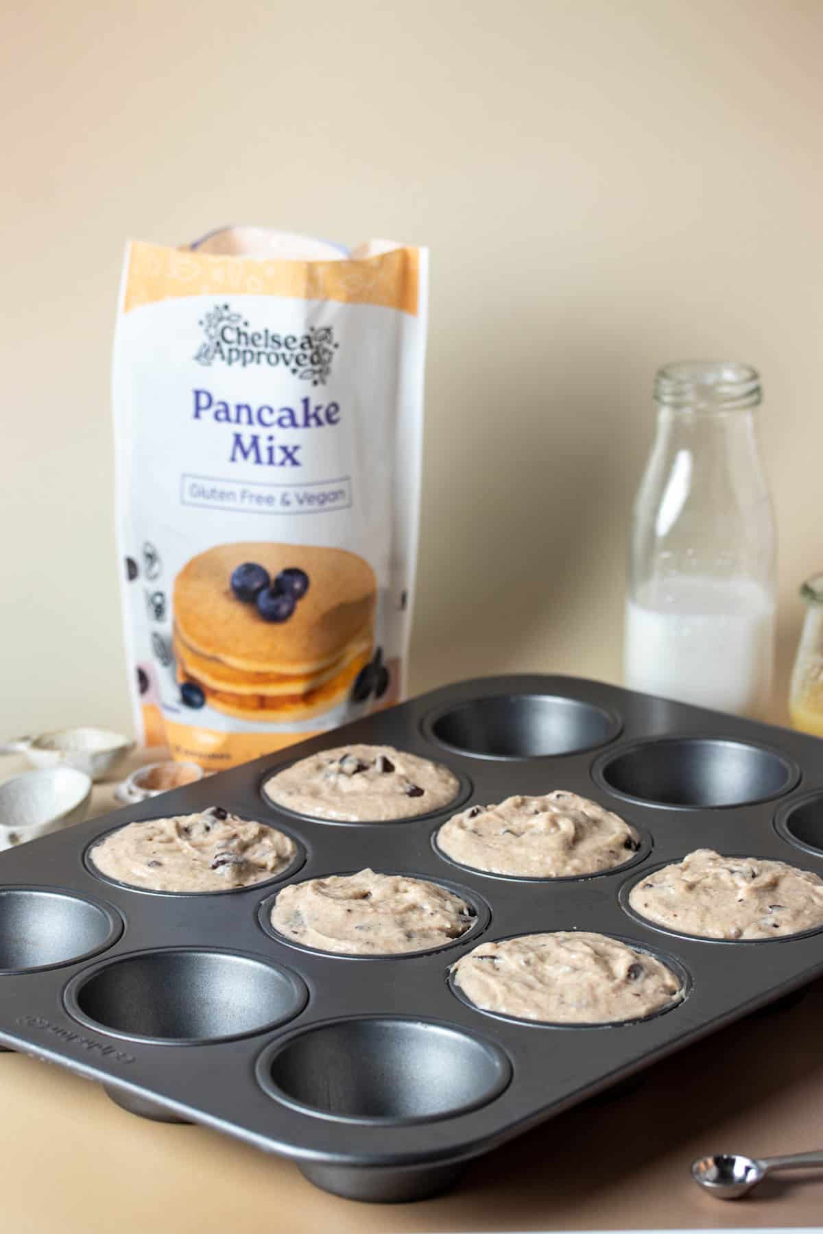 Muffin batter in a muffin tin and ingredients used around it on a tan surface.