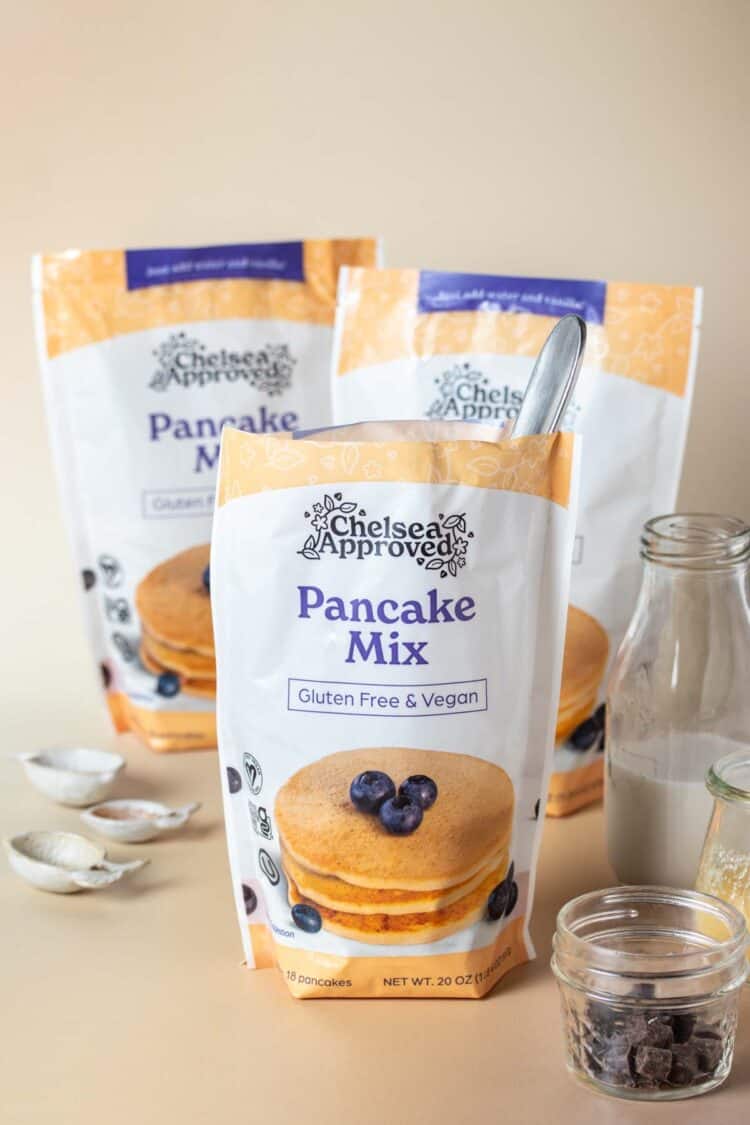 A bag of pancake mix in front of two other bags sitting on a tan surface with various ingredients to make muffins around them.