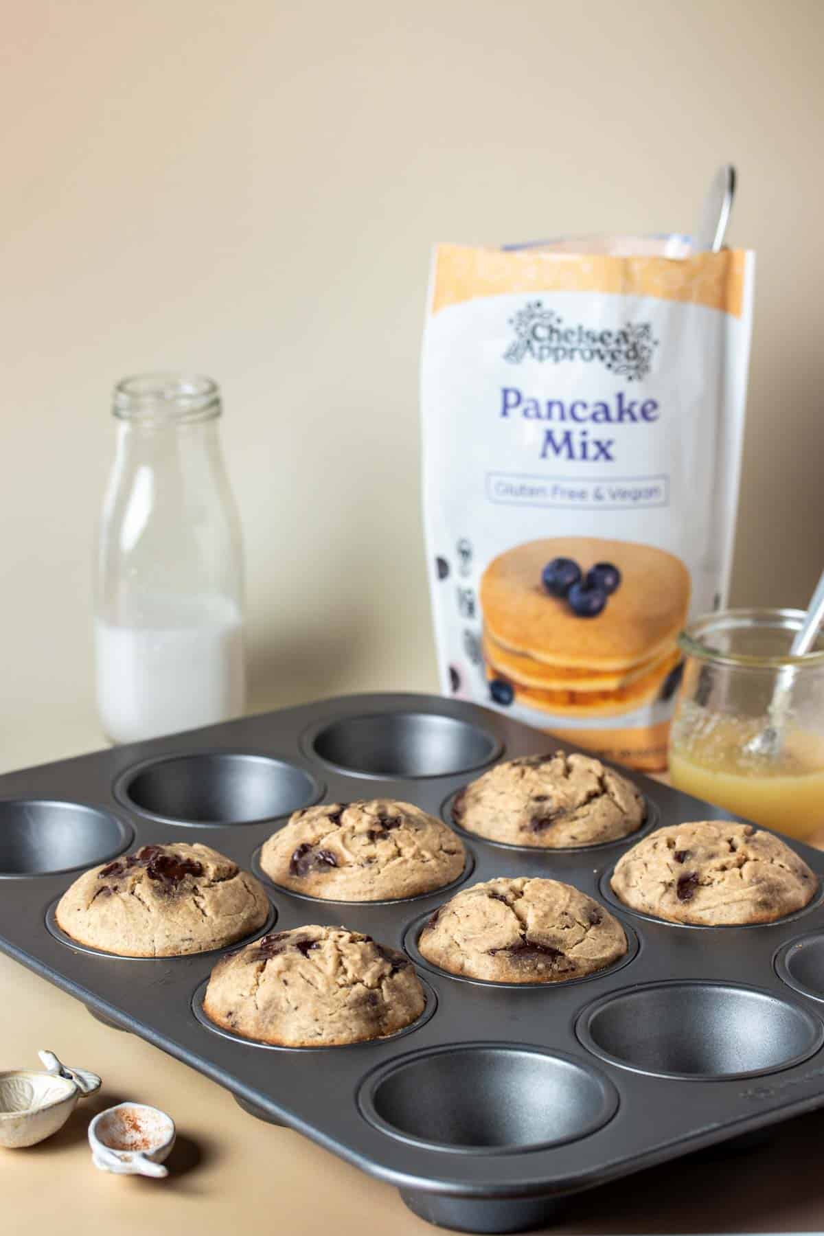 A muffin tin with six baked muffins in it and a bag of pancake mix and baking ingredients around it.