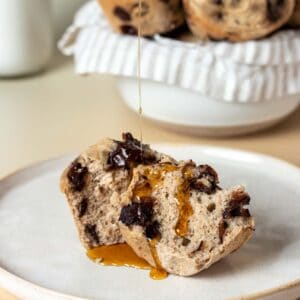 Maple syrup drizzling on a muffin that has been cut in half and laying on a tan plate in front of a bowl of muffins.