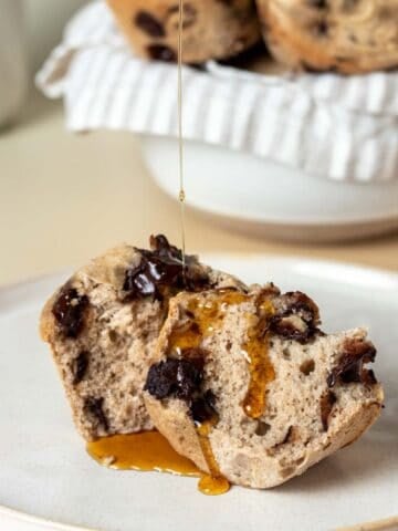 Maple syrup drizzling on a muffin that has been cut in half and laying on a tan plate in front of a bowl of muffins.