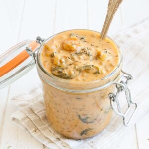 A pink creamy lentil and veggie stew in a glass jar with a hinged top sitting on a white wooden surface.