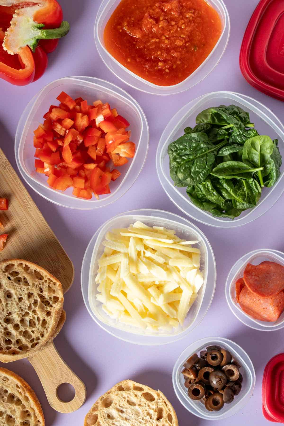 Plastic containers filled with ingredients and toppings for English muffin pizzas and the English muffins next to them.