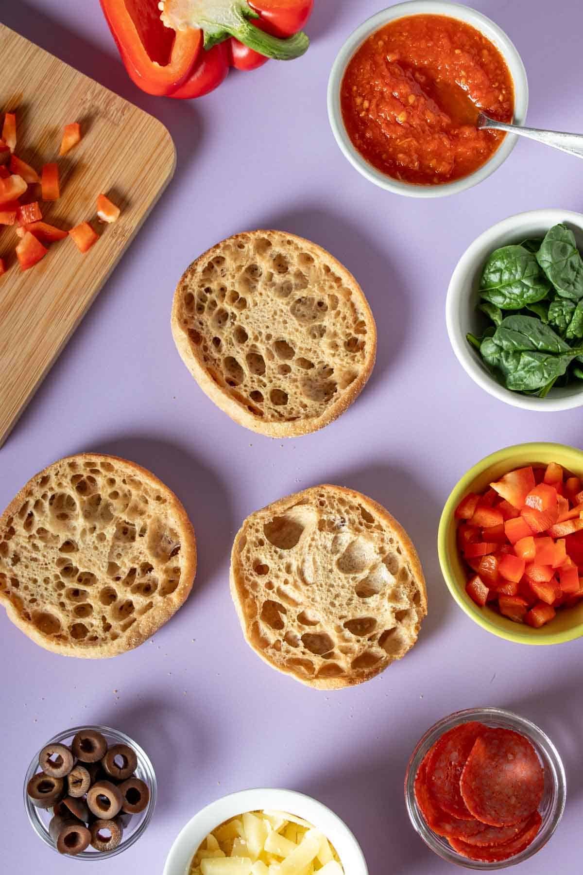 Three English muffins on a purple surface with ingredients for pizzas around them.