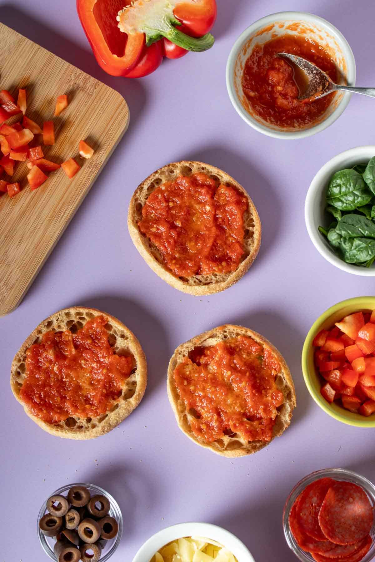 A purple surface with English muffins covered in pizza sauce and toppings around them.