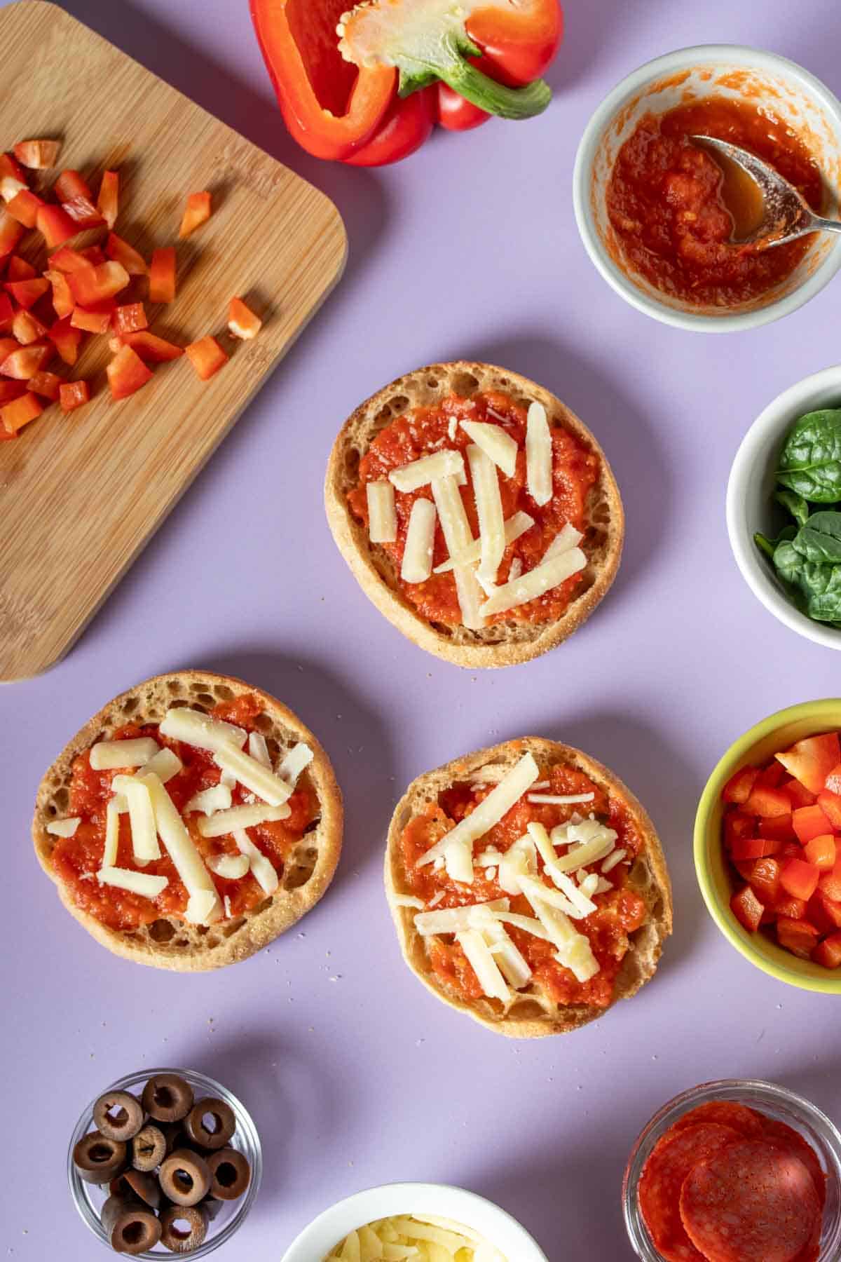 Three English muffins with sauce and cheese and toppings in containers around them on a purple surface.