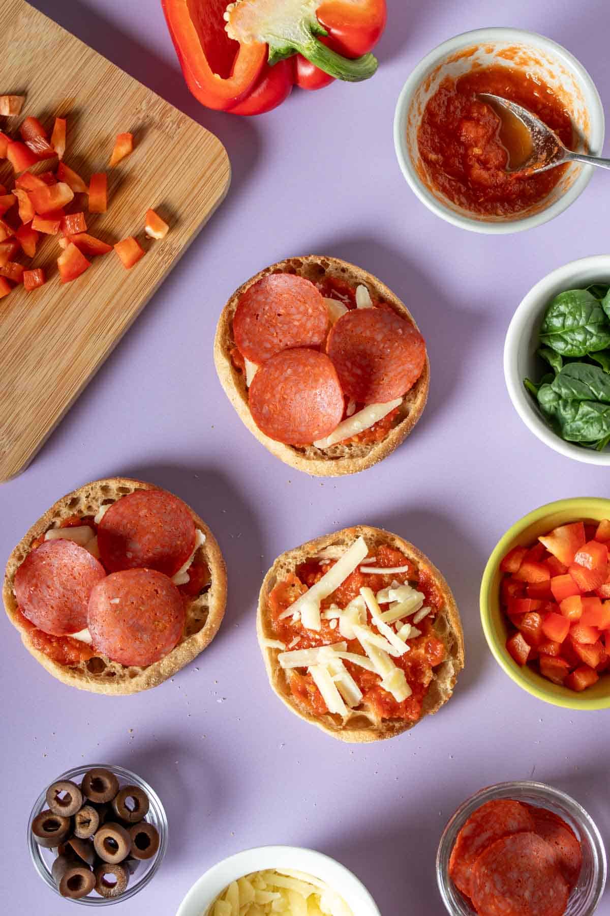 English muffin pizzas being topped with pepperoni and cheese and other ingredients around them on a purple surface.