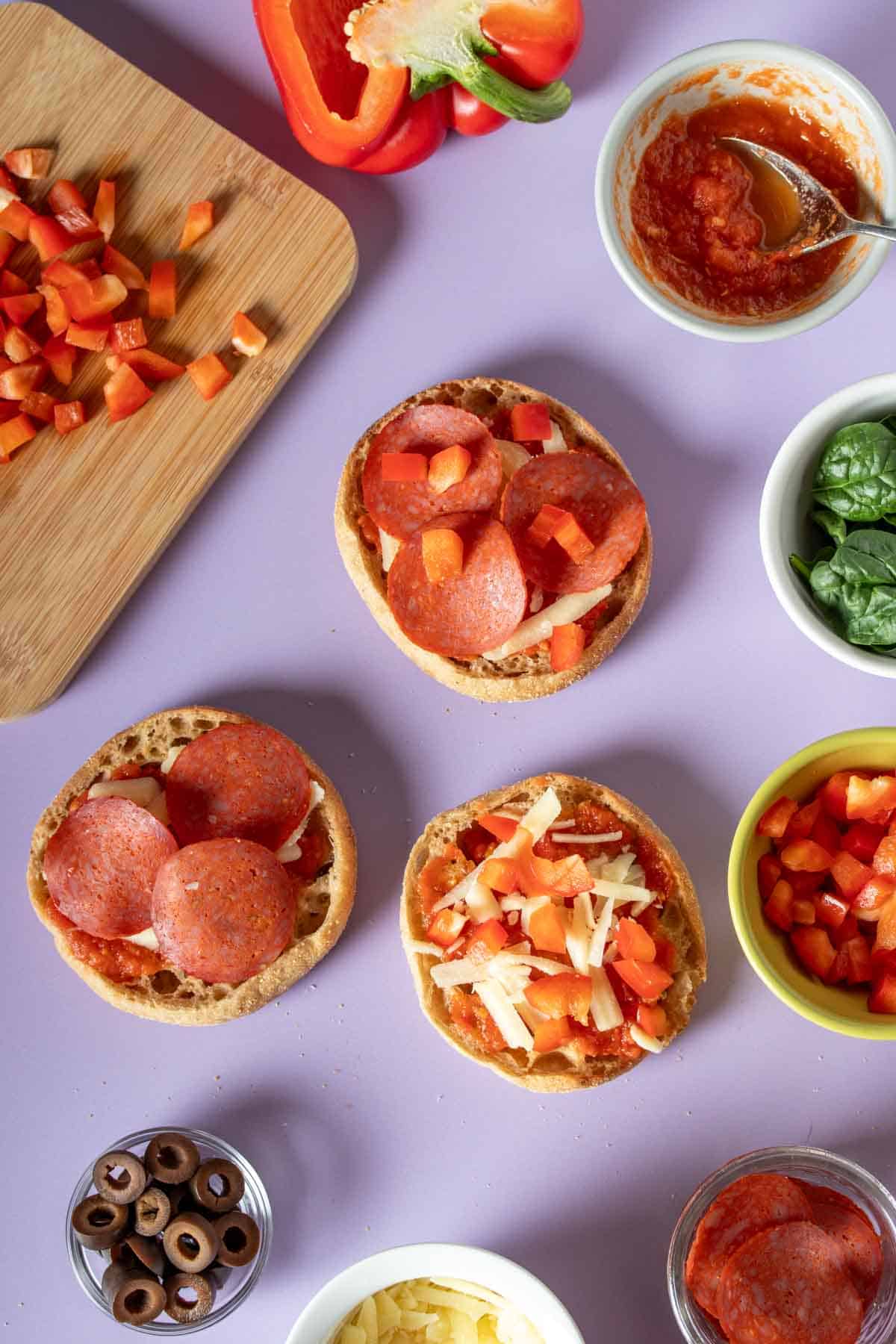 English muffins being made into pizzas sitting on a purple surface with ingredients around them.